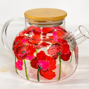 May include: A clear glass teapot featuring a hand-painted design of vibrant red poppy flowers with green stems. The teapot includes a circular light-coloured wooden lid and a glass handle on the side, with a metal coil filter visible inside the spout.