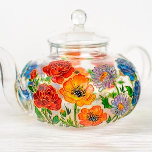 Könnte beinhalten: Eine Teekanne aus klarem Glas mit einem handbemalten Blumenmuster in leuchtendem Rot, Orange und Blau. Die Teekanne verfügt über einen passenden Glasdeckel mit einem kleinen Knauf, einen geschwungenen Ausguss und einen abgerundeten Griff.