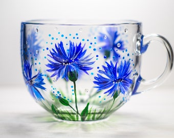 May include: A clear glass teacup with a handle, hand-painted with vibrant blue cornflowers and green stems. The design includes blue dots and a touch of green grass at the base, creating a floral theme.