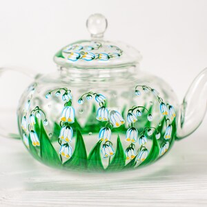 May include: A clear glass teapot with a painted design of white lily of the valley flowers with green leaves and yellow accents.