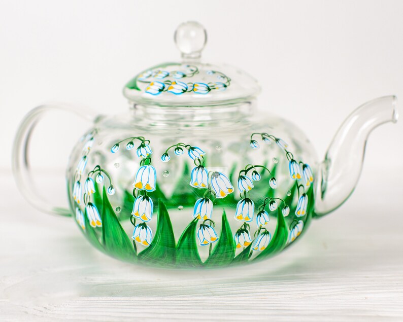 May include: A clear glass teapot with a painted design of white lily of the valley flowers with green leaves and yellow accents.