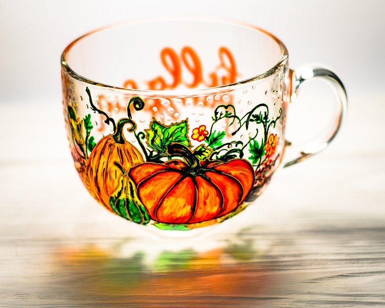 May include: A clear glass mug featuring a hand-painted design of orange pumpkins, green vines, and small yellow flowers. The exterior surface displays a painted orange script text. The mug has a curved handle and a wide, rounded bowl shape.