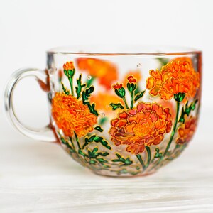 May include: A clear glass teacup with a handle, adorned with hand-painted orange marigold flowers and green leaves. The floral design is outlined in gold, adding a touch of elegance to this decorative teacup.