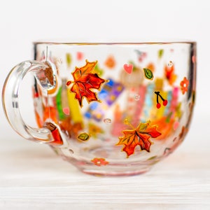 Puede incluir: Una taza de cristal transparente con dise&ntilde;os pintados a mano de hojas de arce oto&ntilde;ales en tonos naranja y rojo, peque&ntilde;os corazones rosas, cerezas rojas y flores diminutas. La taza cuenta con un asa curva y una superficie lisa.