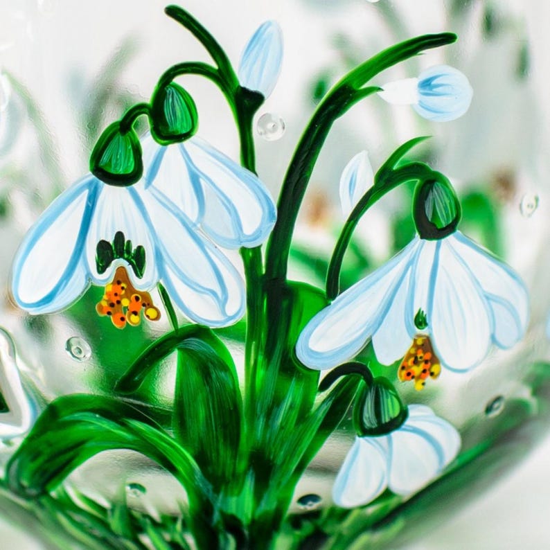 May include: A clear glass surface decorated with hand-painted snowdrop flowers. The design features white petals with light blue accents, vibrant green stems and leaves, and small orange centers. The glass has a textured finish with small bubbles.
