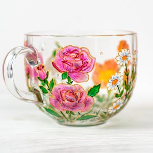 K&ouml;nnte beinhalten: Transparente Teetasse mit gro&szlig;em Henkel, handbemalt mit rosa Rosen, wei&szlig;en G&auml;nsebl&uuml;mchen und orangefarbenen Blumen. Das florale Design ist goldfarben umrandet, mit gr&uuml;nen Bl&auml;ttern und Stielen. Die Tasse steht auf einer wei&szlig;en Oberfl&auml;che.