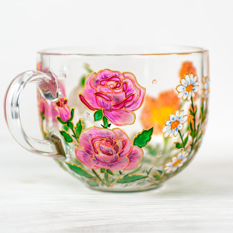 K&ouml;nnte beinhalten: Transparente Teetasse mit gro&szlig;em Henkel, handbemalt mit rosa Rosen, wei&szlig;en G&auml;nsebl&uuml;mchen und orangefarbenen Blumen. Das florale Design ist goldfarben umrandet, mit gr&uuml;nen Bl&auml;ttern und Stielen. Die Tasse steht auf einer wei&szlig;en Oberfl&auml;che.