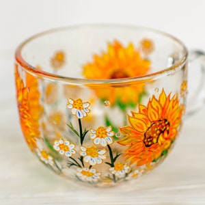 Op de afbeelding: Een doorzichtige glazen mok met handgeschilderde gele zonnebloemen en kleine witte madeliefjes met gele harten. Het bloemmotief loopt rond de buitenkant van de beker, die is voorzien van een glazen handvat aan de rechterkant en een afgeronde rand.