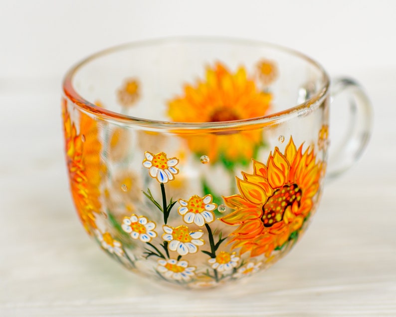 Op de afbeelding: Een doorzichtige glazen mok met handgeschilderde gele zonnebloemen en kleine witte madeliefjes met gele harten. Het bloemmotief loopt rond de buitenkant van de beker, die is voorzien van een glazen handvat aan de rechterkant en een afgeronde rand.