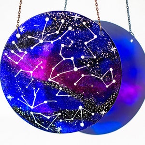 May include: A round glass bauble with a blue and purple galaxy background and white constellations. The bauble is hanging from a gold chain.