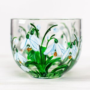 May include: A clear glass bowl featuring a hand-painted design of white snowdrop flowers with green stems and leaves. The floral artwork wraps around the exterior of the bowl, showcasing delicate white petals with yellow centers and green accents.