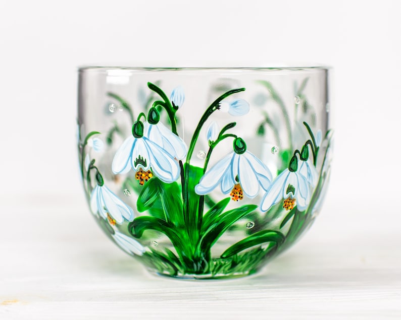 May include: A clear glass bowl featuring a hand-painted design of white snowdrop flowers with green stems and leaves. The floral artwork wraps around the exterior of the bowl, showcasing delicate white petals with yellow centers and green accents.