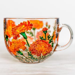 May include: A clear glass teacup with a handle, adorned with hand-painted orange marigold flowers and green stems. The floral design is detailed with red accents and small, clear embellishments. The cup is set against a white background.