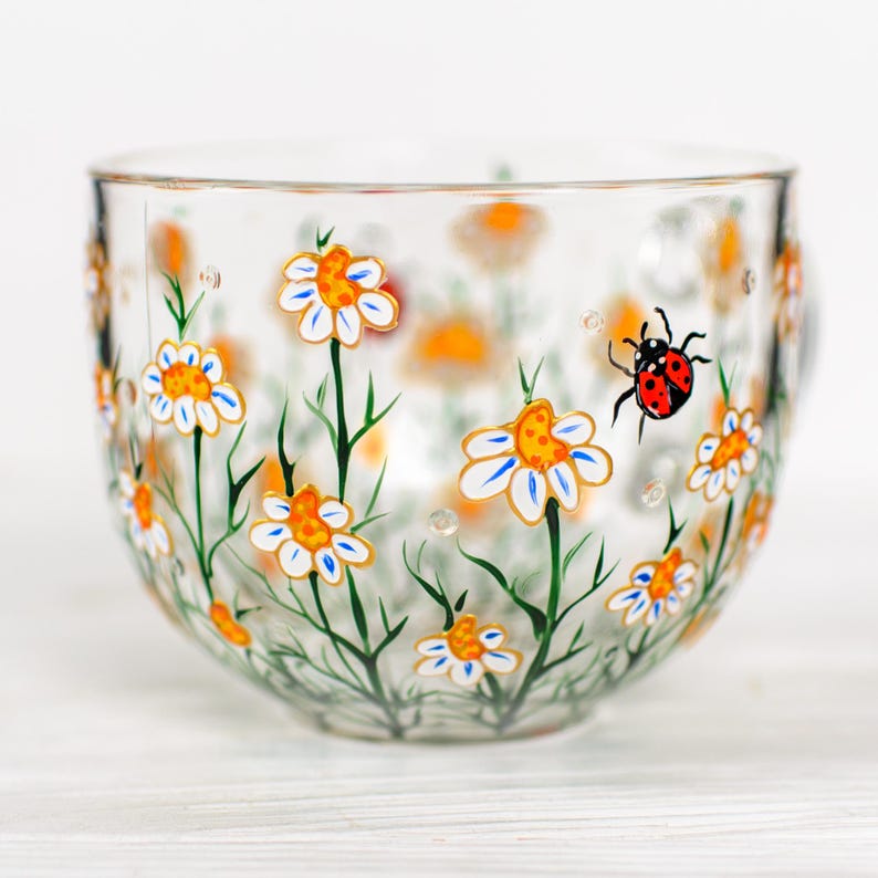 Puede incluir: Una taza de t&eacute; de vidrio transparente con un dise&ntilde;o floral pintado. Las margaritas blancas con centros naranjas y detalles azules est&aacute;n dispersas por la taza, junto con una mariquita roja y negra. Los tallos y hojas verdes completan la escena.