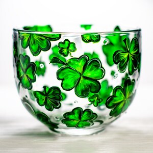 May include: A clear glass bowl decorated with hand-painted green shamrocks. The shamrocks are in various sizes and are evenly spaced around the bowl. The bowl is ideal for serving snacks or as a decorative piece.