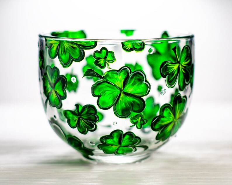 May include: A clear glass bowl decorated with hand-painted green shamrocks. The shamrocks are in various sizes and are evenly spaced around the bowl. The bowl is ideal for serving snacks or as a decorative piece.