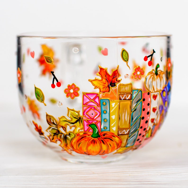 Puede incluir: Una taza de cristal transparente con dise&ntilde;os pintados a mano de libros apilados de colores, calabazas naranjas, hojas de oto&ntilde;o, peque&ntilde;as cerezas rojas y flores diminutas. Las ilustraciones vibrantes decoran el exterior del recipiente.