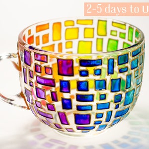 May include: A clear glass mug featuring a colorful stained glass effect with hand-painted rectangular shapes in shades of red, orange, yellow, green, blue, and purple, outlined in gold. The mug has a curved handle and a smooth, transparent finish.