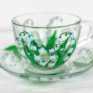 May include: A clear glass teacup and saucer set. The cup and saucer are decorated with hand-painted green leaves and blue and white lily of the valley flowers. The cup has a handle and sits on a matching saucer.