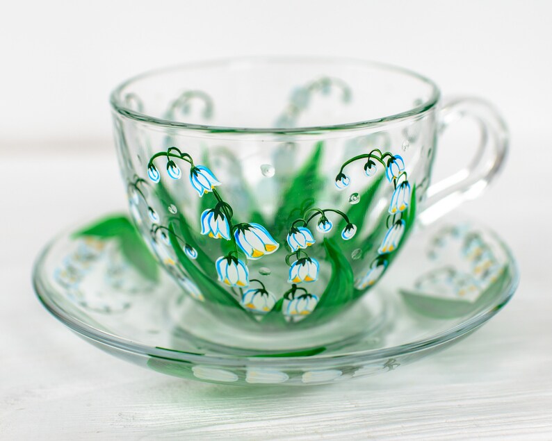 May include: A clear glass teacup and saucer set. The cup and saucer are decorated with hand-painted green leaves and blue and white lily of the valley flowers. The cup has a handle and sits on a matching saucer.