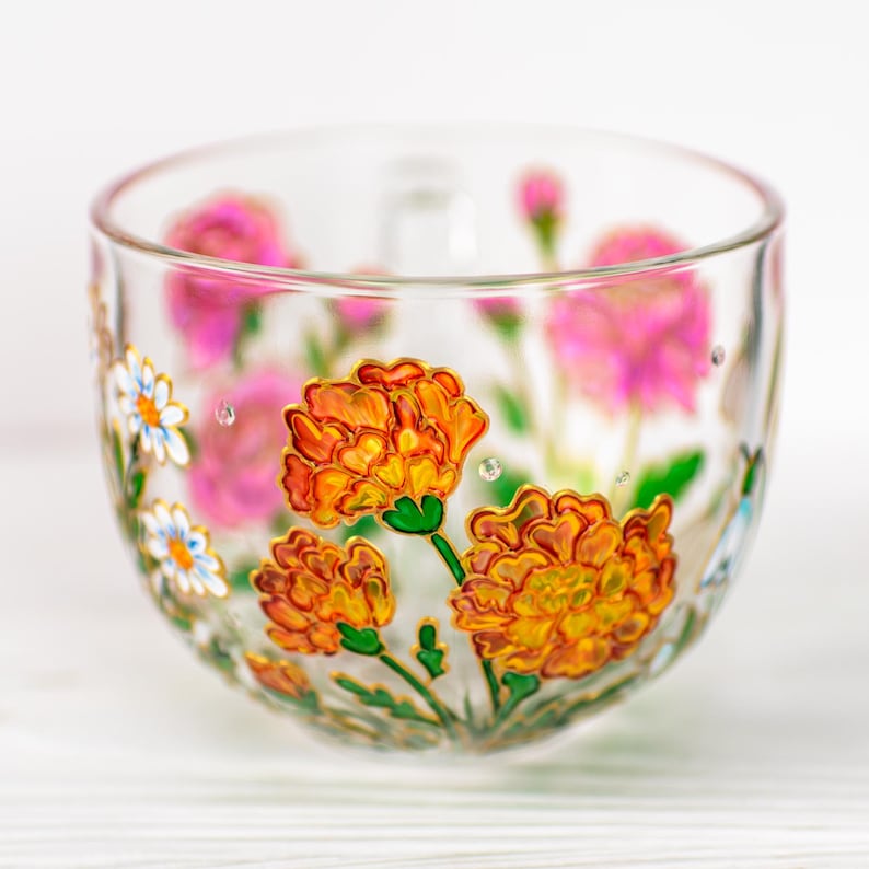 K&ouml;nnte beinhalten: Eine transparente Teetasse aus Glas mit handgemalten Blumenmustern. Die Tasse zeigt orange und gelbe Ringelblumen mit gr&uuml;nen Stielen und Bl&auml;ttern sowie rosa Bl&uuml;ten. Die Blumen sind goldfarben umrandet. Kleine wei&szlig;e G&auml;nsebl&uuml;mchen und klare Kristalle sind ebenfalls auf die Tasse gemalt.