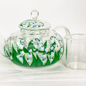 May include: A clear glass teapot with a painted design of white lily of the valley flowers and green leaves. The teapot has a removable strainer and a lid.