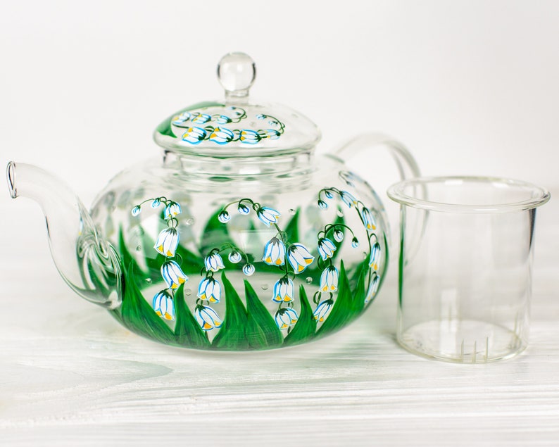 May include: A clear glass teapot with a painted design of white lily of the valley flowers and green leaves. The teapot has a removable strainer and a lid.