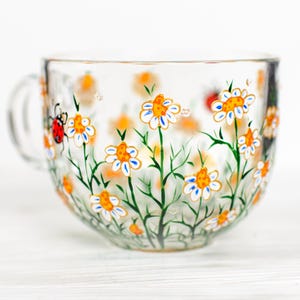 Puede incluir: Una taza de t&eacute; de vidrio transparente con asa, pintada a mano con un dise&ntilde;o floral. Las margaritas blancas con centros naranjas y detalles azules est&aacute;n dispersas por la taza, junto con tallos y hojas verdes. Una mariquita roja tambi&eacute;n est&aacute; pintada en la taza.