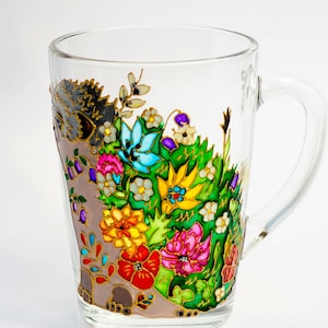 May include: A glass mug with a hand-painted design of a hedgehog surrounded by colorful flowers. The flowers are painted in shades of red, yellow, blue, green, and purple.
