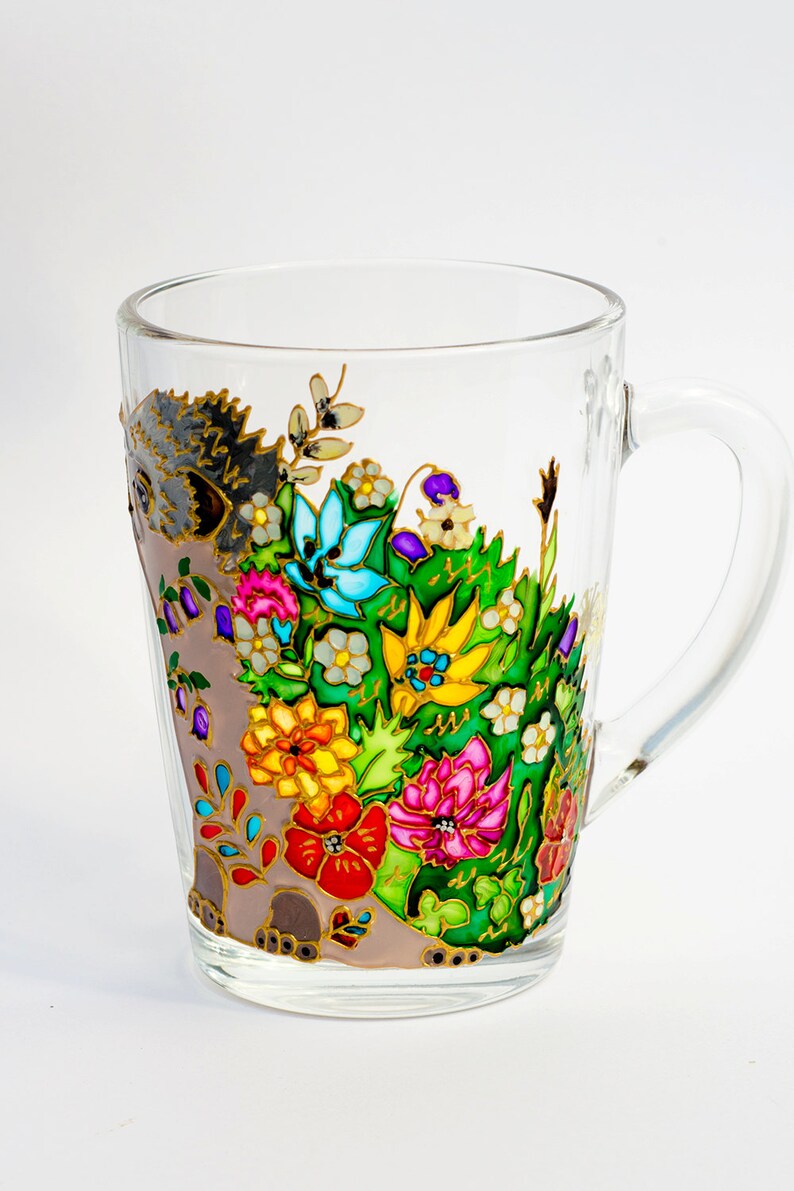 May include: A glass mug with a hand-painted design of a hedgehog surrounded by colorful flowers. The flowers are painted in shades of red, yellow, blue, green, and purple.