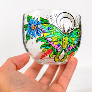 May include: A clear glass bowl with a hand-painted design featuring a colorful butterfly, blue flower, and foliage. The butterfly has green, yellow, and blue wings. The bowl is held in a hand.