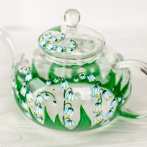 May include: A clear glass teapot with a painted design of white and blue lily of the valley flowers on a green stem. The teapot has a lid and a handle.