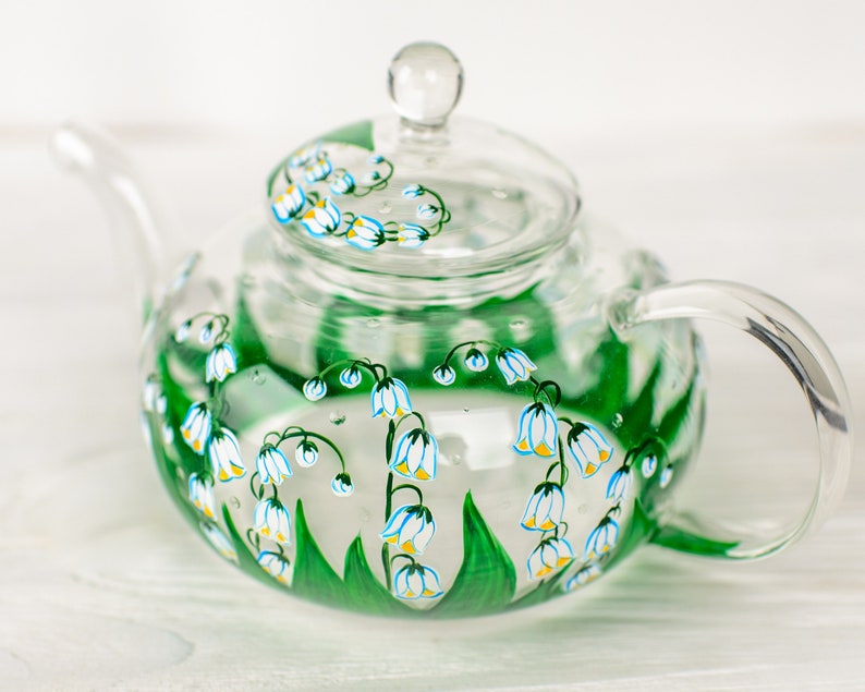 May include: A clear glass teapot with a painted design of white and blue lily of the valley flowers on a green stem. The teapot has a lid and a handle.