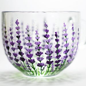 May include: A clear glass mug featuring a hand-painted design of multiple lavender stalks in various shades of purple and green. The floral pattern wraps around the exterior of the cup, showcasing detailed petals and stems against the transparent glass.