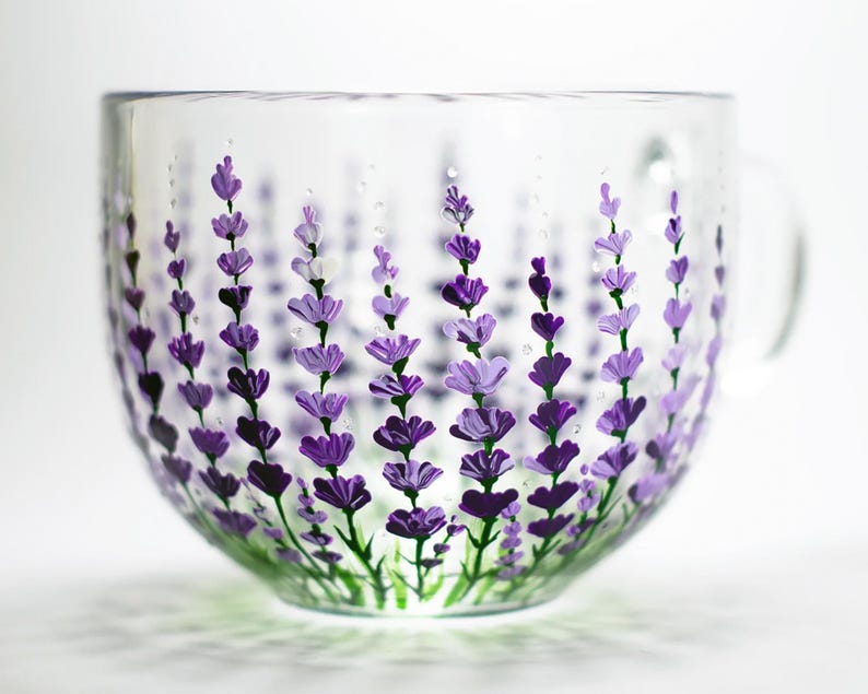 May include: A clear glass mug featuring a hand-painted design of multiple lavender stalks in various shades of purple and green. The floral pattern wraps around the exterior of the cup, showcasing detailed petals and stems against the transparent glass.