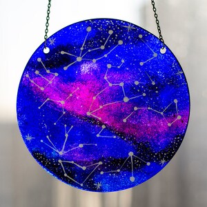 May include: A round glass bauble with a blue and pink galaxy design and white constellations. The bauble is hanging from a silver chain.