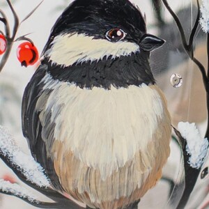 May include: A hand-painted glass ornament featuring a black-capped chickadee with a black head, white cheeks, and a tan-colored breast. The bird is perched on a dark branch dusted with white snow, accented by bright red berries and small clear gems.