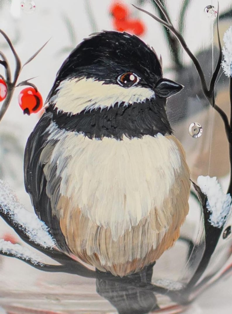May include: A hand-painted glass ornament featuring a black-capped chickadee with a black head, white cheeks, and a tan-colored breast. The bird is perched on a dark branch dusted with white snow, accented by bright red berries and small clear gems.