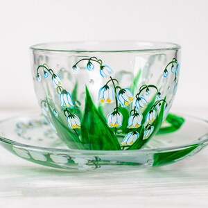 May include: A clear glass teacup and saucer set. The cup and saucer are decorated with hand-painted lily of the valley flowers. The flowers have white petals with yellow centers and are accented with green leaves and stems. The handle is green.