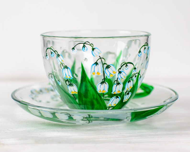 May include: A clear glass teacup and saucer set. The cup and saucer are decorated with hand-painted lily of the valley flowers. The flowers have white petals with yellow centers and are accented with green leaves and stems. The handle is green.