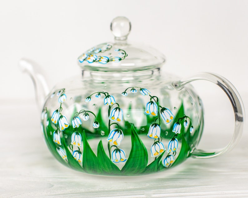May include: A clear glass teapot with a lid, hand-painted with a floral design of white lily of the valley flowers and green leaves.