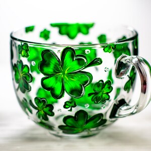 May include: A clear glass teacup with a large handle, decorated with vibrant green shamrocks. The shamrocks are painted on the outside of the cup, creating a festive and cheerful design. The cup is perfect for St. Patrick's Day.
