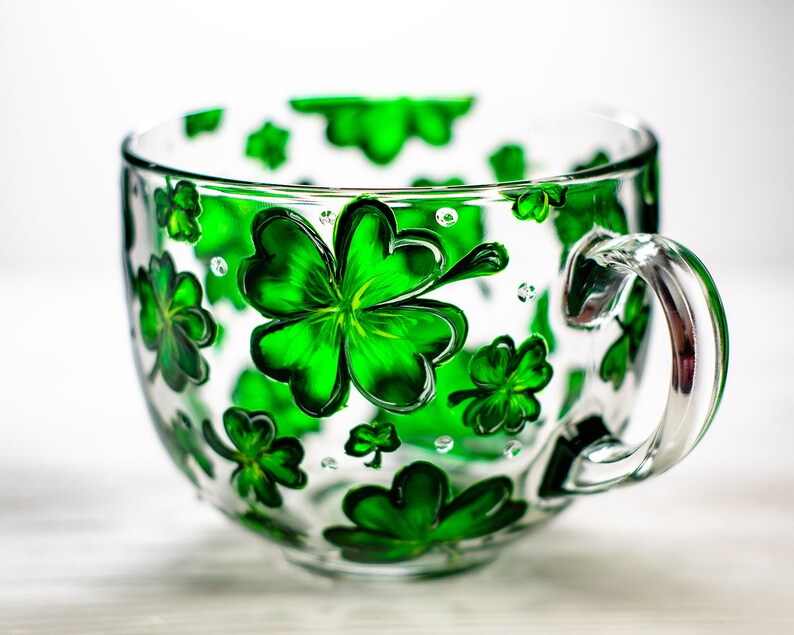 May include: A clear glass teacup with a large handle, decorated with vibrant green shamrocks. The shamrocks are painted on the outside of the cup, creating a festive and cheerful design. The cup is perfect for St. Patrick's Day.