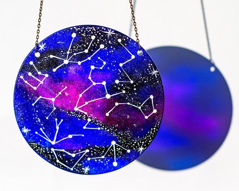 May include: A round glass bauble with a blue and purple galaxy background and white constellations painted on it. The bauble is hanging from a gold chain.
