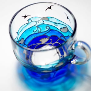 Puede incluir: Una taza de t&eacute; de vidrio transparente con un dise&ntilde;o de olas oce&aacute;nicas pintado a mano en tonos azules y grises. La taza tiene un asa y est&aacute; decorada con tres siluetas de p&aacute;jaros negros. El dise&ntilde;o de la taza es visible desde dentro y fuera.