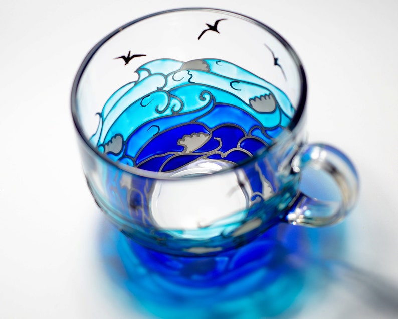 Puede incluir: Una taza de t&eacute; de vidrio transparente con un dise&ntilde;o de olas oce&aacute;nicas pintado a mano en tonos azules y grises. La taza tiene un asa y est&aacute; decorada con tres siluetas de p&aacute;jaros negros. El dise&ntilde;o de la taza es visible desde dentro y fuera.