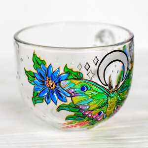 May include: A clear glass bowl with a hand-painted design featuring a blue flower and a green and yellow butterfly. The bowl has a rounded shape and is decorated with gold accents and small diamond shapes.