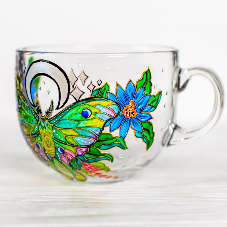May include: A clear glass mug with a hand-painted design featuring a colorful butterfly, a blue flower, and a crescent moon. The butterfly has green, yellow, and blue wings. The flower has blue petals and an orange center. The mug has a handle.
