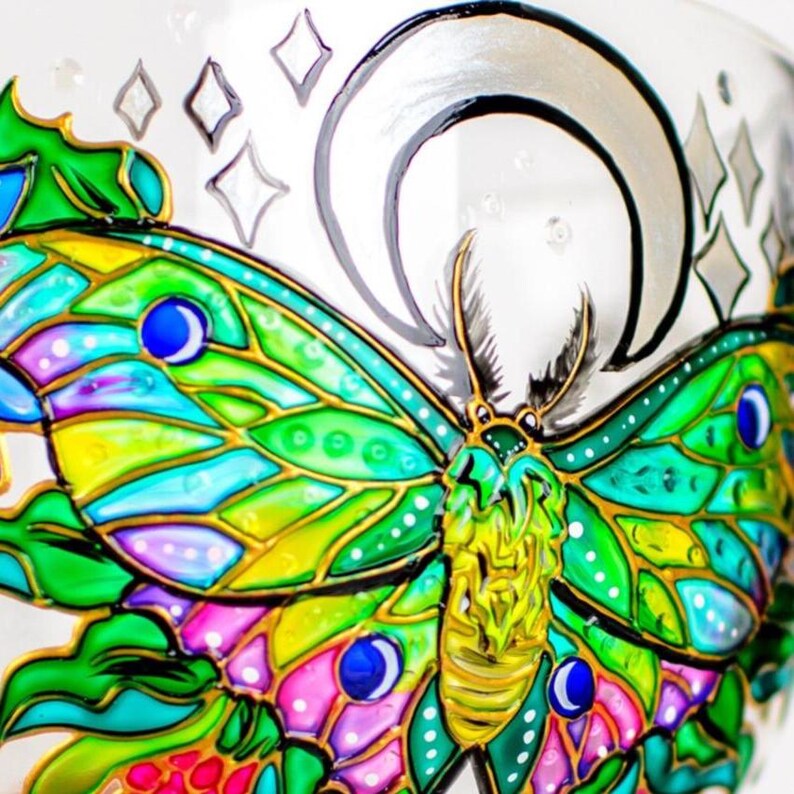 May include: A clear glass surface featuring a hand-painted moth with vibrant green, yellow, and pink wings outlined in gold. The moth is positioned beneath a silver crescent moon and surrounded by small, outlined diamond-shaped star symbols.