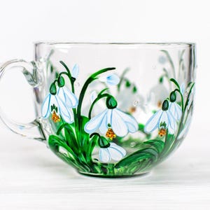 Puede incluir: Una taza de t&eacute; de vidrio transparente con asa, pintada a mano con flores blancas de campanillas de invierno, tallos y hojas verdes. Las flores tienen centros amarillos. La taza est&aacute; sobre una superficie blanca.